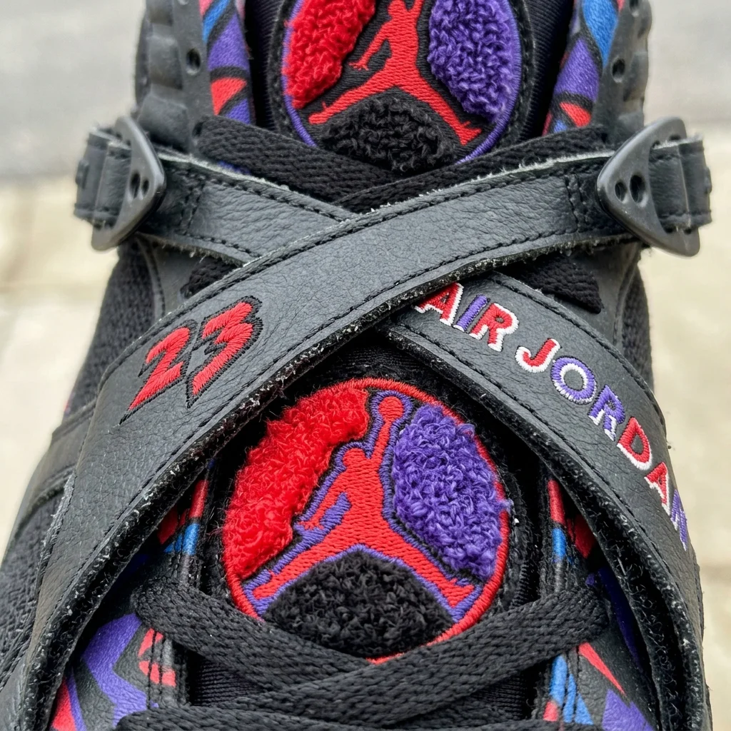 Close-up macro photography of the crossing midfoot straps and chenille tongue logo of an Air Jordan 8 sneaker, highlighting the maximalist 1990s design, photorealistic.